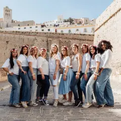 Dix amies célèbrent leur evjf en France, souriantes et parées de couronnes de fleurs, devant un mur de pierre et des bâtiments blancs.