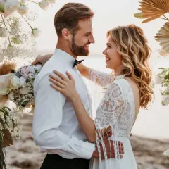 Un couple rayonnant s'embrasse sous une arche fleurie, célébrant leur mariage sans enfant sur une plage.