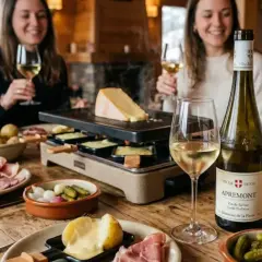 Un groupe d'amis savoure une raclette conviviale, accompagnée de pommes de terre, charcuterie et cornichons. Une bouteille de vin blanc de Savoie, parfait pour accompagner ce plat, trône sur la table en bois.