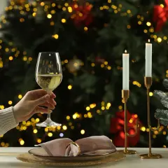 Une main tient un verre de vin blanc pétillant, parfait pour un buffet froid. Une bouteille décorée et des bougies illuminent la scène devant un sapin de Noël scintillant.