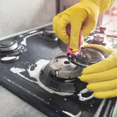 Mains jaunes pour nettoyer graisse brûlée gazinière. Une éponge rose frotte une partie du brûleur.