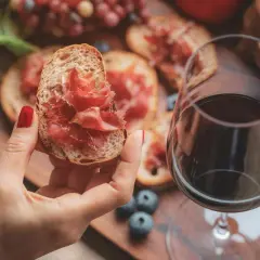 Une main tient une tartine garnie de jambon cru, à côté d'un verre de vin rouge. Parfait pour un apéro dinatoire.