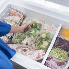 Une main range des légumes et fruits congelés dans un congélateur. On peut congeler un plat cuisiné pour le conserver.