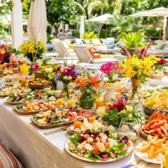 Un grand buffet en plein air, décoré de fleurs colorées, invite à un repas entre amis. Des fruits de mer, fromages et salades composent ce festin estival.