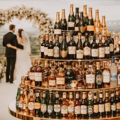 Une pyramide de bouteilles pour un vin d'honneur de mariage. Les mariés se tiennent sous une arche fleurie.