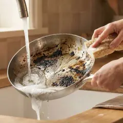 Nettoyer poêle bicarbonate vinaigre : une main frotte une poêle brûlée sous l'eau courante avec une éponge.