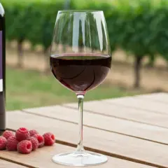 Verre de Pinot Noir, framboises et bouteille sur une table en bois, avec des vignes en arrière-plan. La température idéale pour ce vin est de 14-16°C.