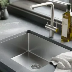 Un évier inox rayé, prêt à être nettoyé. Une bouteille d'huile d'olive et un torchon à proximité.