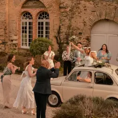 Mariage Dolce Vita : la mariée, radieuse, lève son bouquet depuis une Fiat 500. Ses invités applaudissent devant un manoir en pierre.