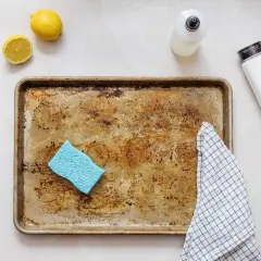 Une plaque de cuisson en aluminium très sale, une éponge bleue, des citrons, une bouteille de produit vaisselle et une serviette à carreaux, prêts à décaper l'aluminium.