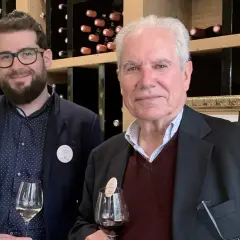 Deux hommes dégustent du vin devant des étagères remplies de bouteilles. L'un porte un badge "Domaine Rozel - Grignan-les-Adhémar avis".