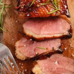 Magret de canard rosé, tranché sur une planche en bois, garni de romarin. Une fourchette et une gousse d'ail sont à proximité. Un délice pour accompagner un bon vin.