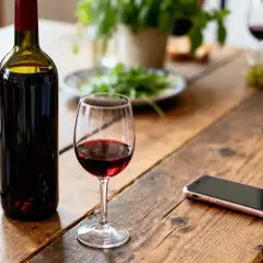 Bouteille de vin rouge et verre sur une table en bois, prêts pour une fondue vigneronne. Un téléphone portable est branché à proximité.