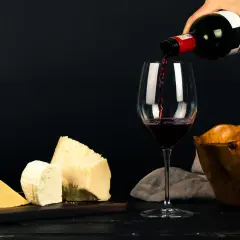 Un verre se remplit de vin rouge, à côté de morceaux de fromage et d'un bol en bois.