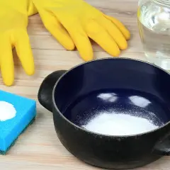 Gants jaunes, éponge bleue avec poudre blanche, casserole noire et un bocal rempli de liquide. Idéal pour savoir comment enlever le cramer dans une casserole sans bicarbonate.