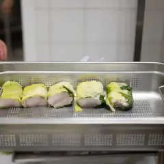 Cinq rouleaux de poisson enroulés dans des feuilles de chou, prêts pour la cuisson. Une main est visible en arrière-plan, préparant ce plat pour la journée internationale des cuisiniers.