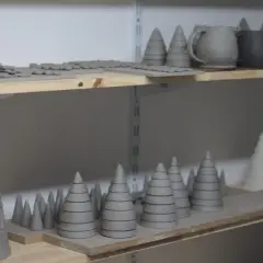 Sur des étagères, des pièces de céramique brutes en forme de sapins et de pots attendent d'aller au four.
