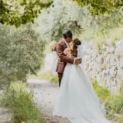 Un couple s'embrasse tendrement lors de leur mariage de 50 personnes, entouré par la nature verdoyante et un mur de pierres anciennes.