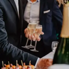 Des hommes en costume portent des flûtes de champagne. Une bouteille de champagne est au premier plan, suggérant un vin d'honneur original.