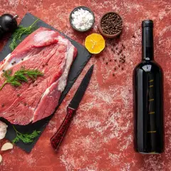 Un morceau de viande crue, prêt pour un repas savoureux avec un vin rouge. Des épices, de l'ail et une tranche de citron complètent la scène.