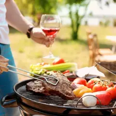 Un homme savoure un verre de vin rouge tout en préparant un barbecue avec des steaks et des légumes.
