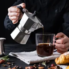 Cafetière italienne inox versant du café dans une tasse. Un petit pot en métal et des croissants sont sur la table.