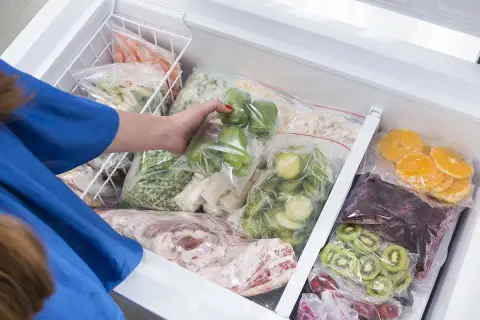 Une main range des légumes et fruits congelés dans un congélateur. On peut congeler un plat cuisiné pour le conserver.