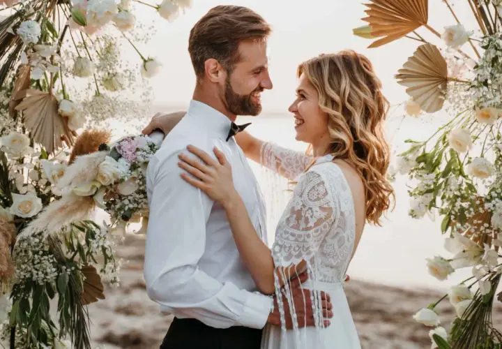 Un couple rayonnant s'embrasse sous une arche fleurie, célébrant leur mariage sans enfant sur une plage.