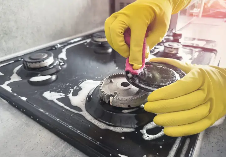 Mains jaunes pour nettoyer graisse brûlée gazinière. Une éponge rose frotte une partie du brûleur.