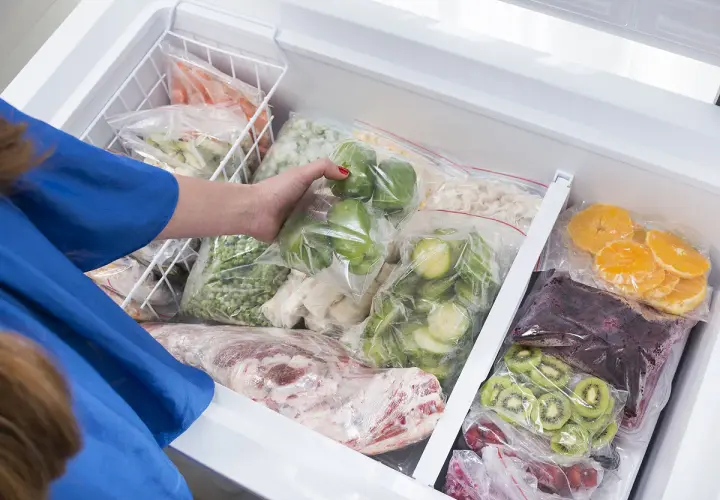 Une main range des légumes et fruits congelés dans un congélateur. On peut congeler un plat cuisiné pour le conserver.