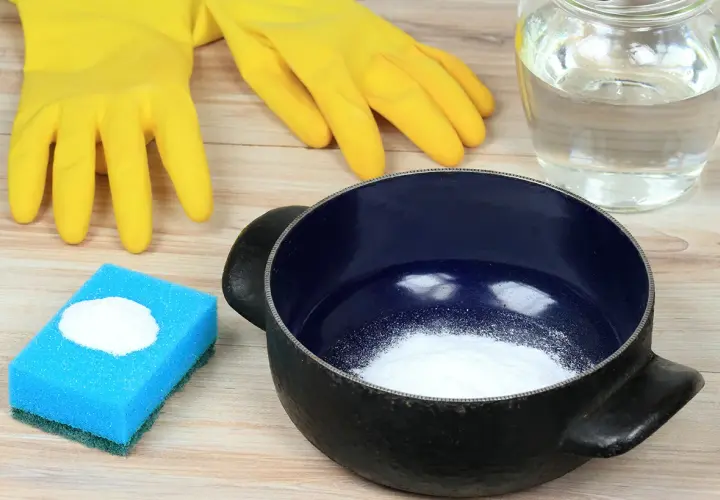 Gants jaunes, éponge bleue avec poudre blanche, casserole noire et un bocal rempli de liquide. Idéal pour savoir comment enlever le cramer dans une casserole sans bicarbonate.