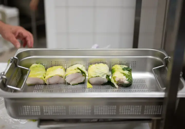 Cinq rouleaux de poisson enroulés dans des feuilles de chou, prêts pour la cuisson. Une main est visible en arrière-plan, préparant ce plat pour la journée internationale des cuisiniers.