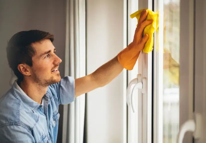 Un homme souriant nettoie une fenêtre en aluminium blanc avec un chiffon jaune.