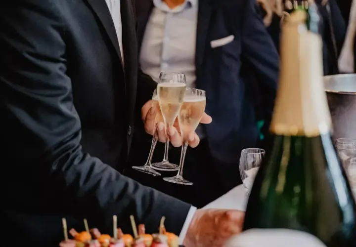 Des hommes en costume portent des flûtes de champagne. Une bouteille de champagne est au premier plan, suggérant un vin d'honneur original.
