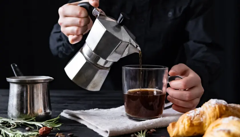 Cafetière italienne inox versant du café dans une tasse. Un petit pot en métal et des croissants sont sur la table.