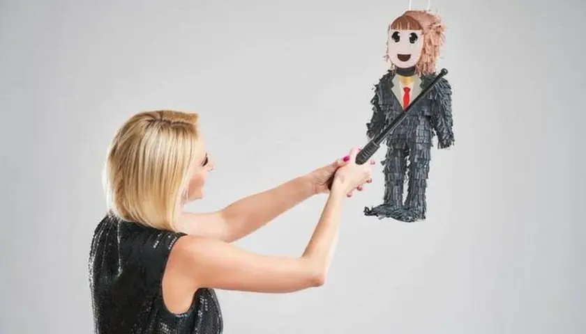 Une femme en robe à paillettes s'apprête à frapper une piñata en forme d'homme en costume, symbole d'une fête de divorce réussie.