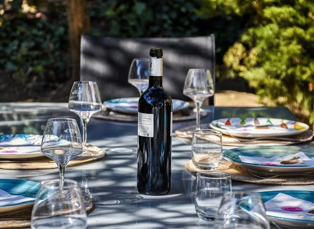 Une bouteille de vin rouge est prête pour un apéro dinatoire en extérieur, entourée de verres et d'assiettes sur une table de jardin.