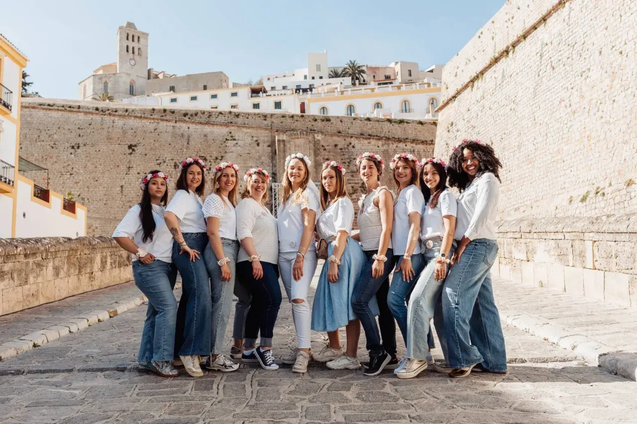 Dix amies célèbrent leur evjf en France, souriantes et parées de couronnes de fleurs, devant un mur de pierre et des bâtiments blancs.
