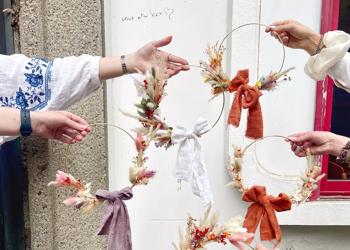 Des mains créatives assemblent des couronnes de fleurs séchées lors d'un atelier EVJF. Ambiance bohème et festive.