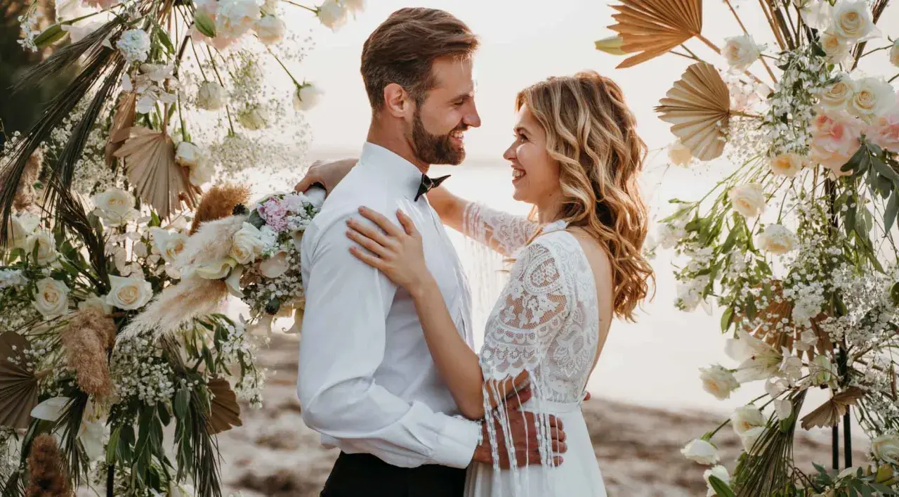 Un couple rayonnant s'embrasse sous une arche fleurie, célébrant leur mariage sans enfant sur une plage.