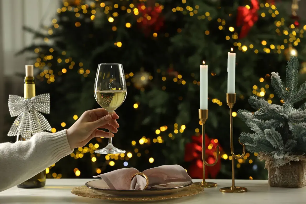 Une main tient un verre de vin blanc pétillant, parfait pour un buffet froid. Une bouteille décorée et des bougies illuminent la scène devant un sapin de Noël scintillant.