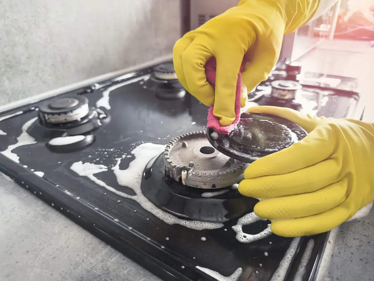 Mains jaunes pour nettoyer graisse brûlée gazinière. Une éponge rose frotte une partie du brûleur.