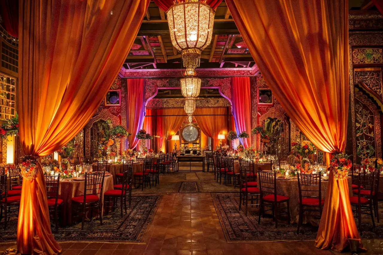 Décoration somptueuse pour un mariage bollywood, avec des drapés orangés, des lustres imposants et des tables dressées dans une ambiance chaleureuse et festive.
