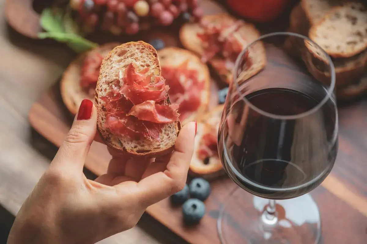 Une main tient une tartine garnie de jambon cru, à côté d'un verre de vin rouge. Parfait pour un apéro dinatoire.