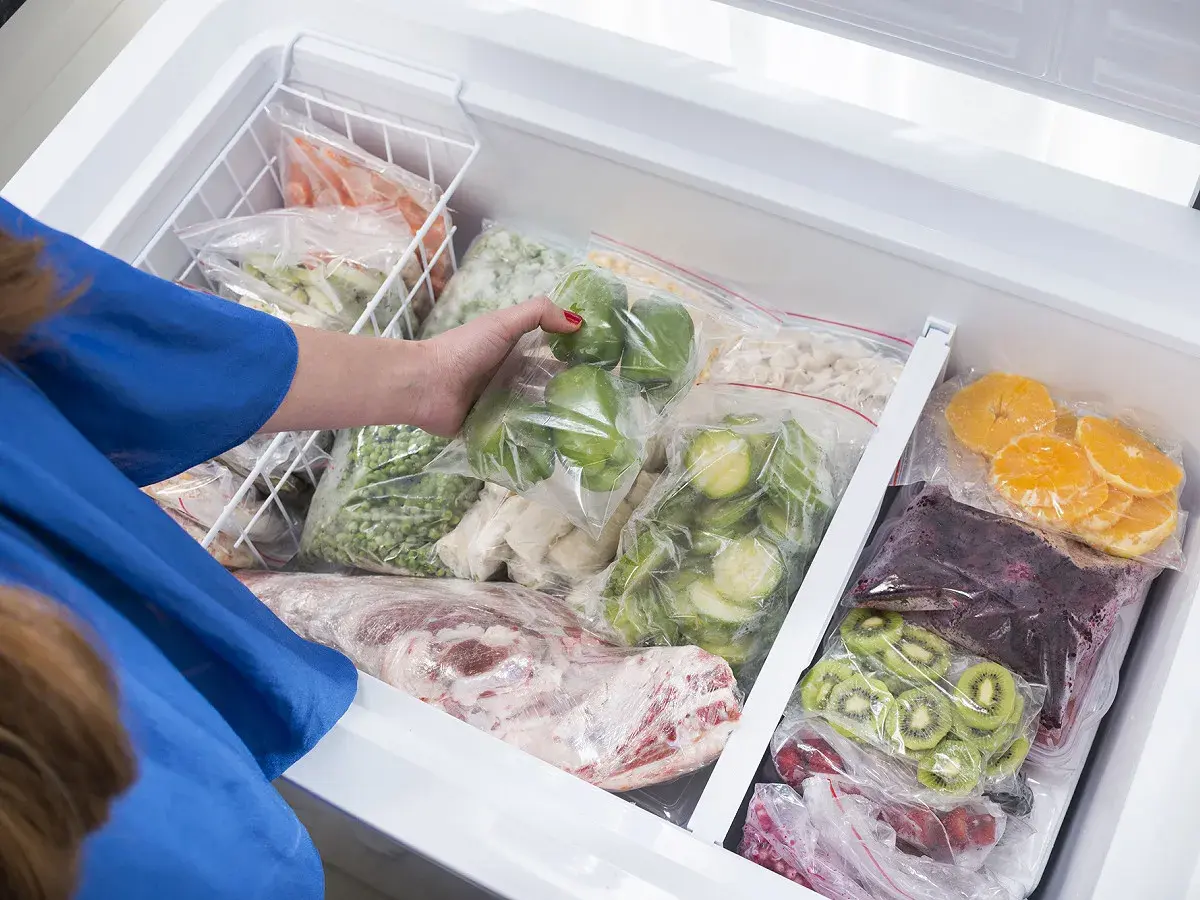 Une main range des légumes et fruits congelés dans un congélateur. On peut congeler un plat cuisiné pour le conserver.