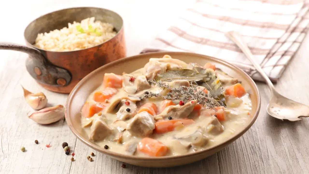 Un plat de blanquette de veau crémeuse avec des carottes et des champignons, servi avec du riz. Un verre de vin rouge est suggéré pour accompagner ce délice.