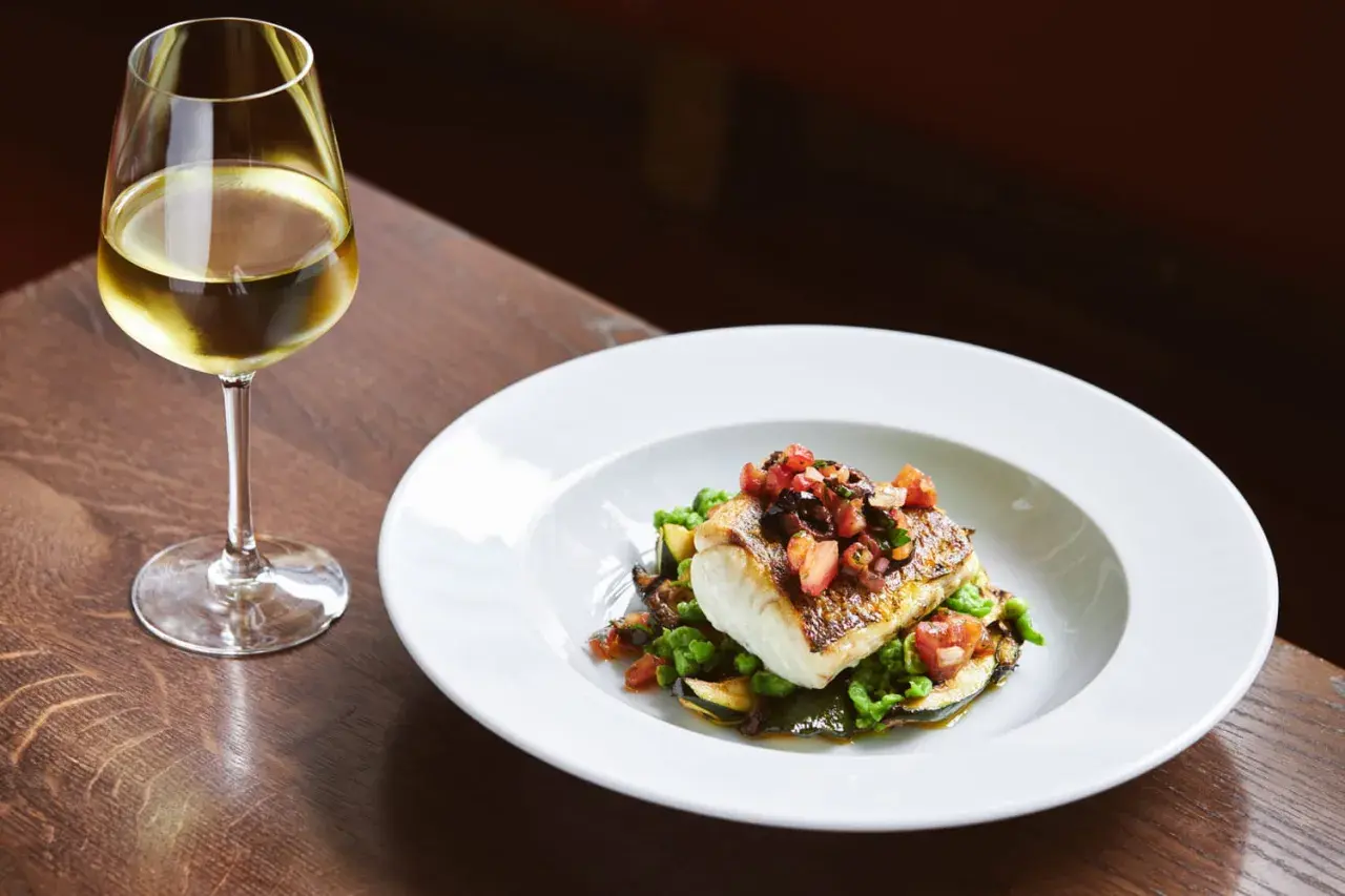 Un verre de vin blanc accompagne un poisson en sauce, servi sur un lit de légumes verts et de courgettes.