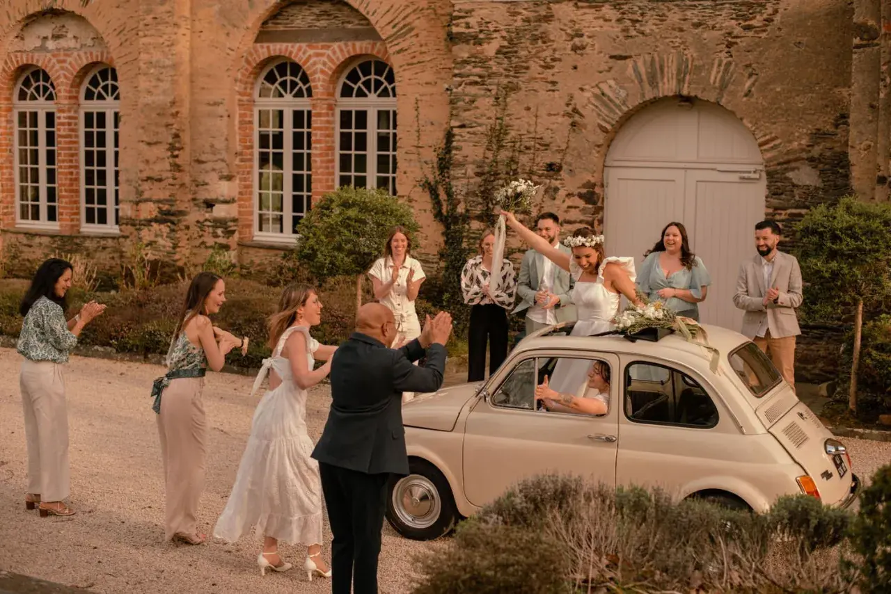 Mariage Dolce Vita : la mariée, radieuse, lève son bouquet depuis une Fiat 500. Ses invités applaudissent devant un manoir en pierre.
