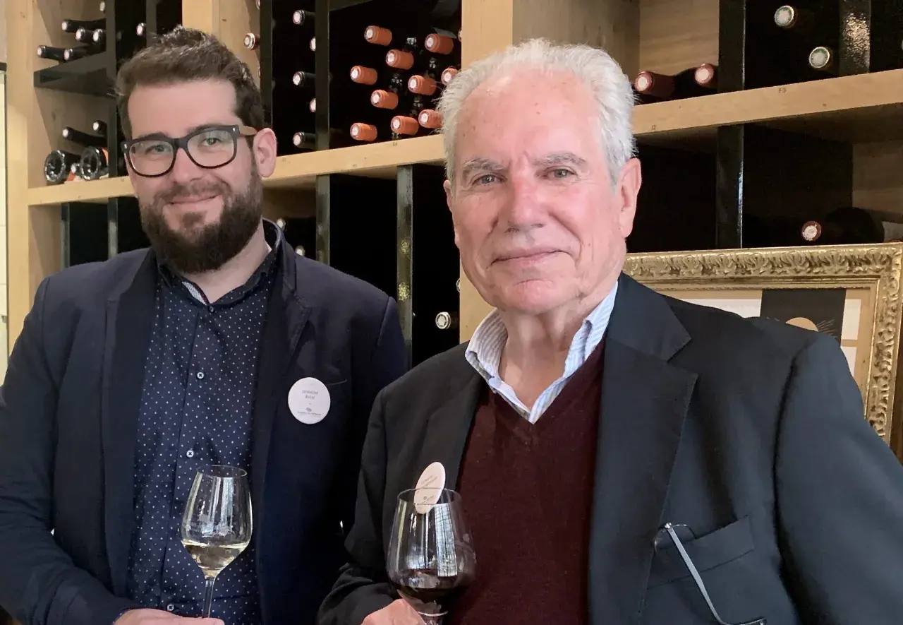 Deux hommes dégustent du vin devant des étagères remplies de bouteilles. L'un porte un badge "Domaine Rozel - Grignan-les-Adhémar avis".