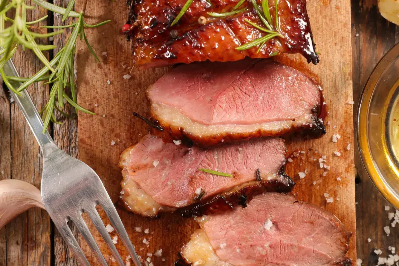 Magret de canard rosé, tranché sur une planche en bois, garni de romarin. Une fourchette et une gousse d'ail sont à proximité. Un délice pour accompagner un bon vin.